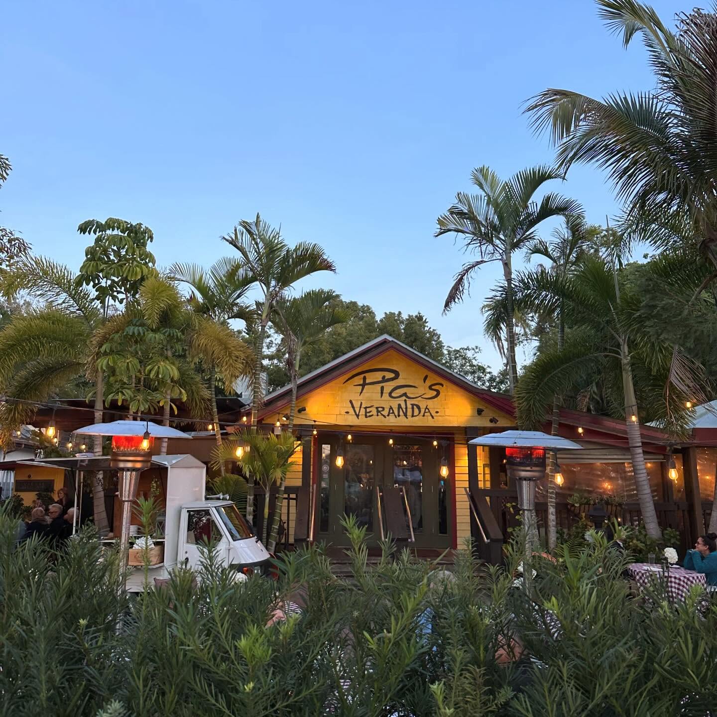 Pia's Veranda at dusk behind a garden of shrubs, patio heaters and palms