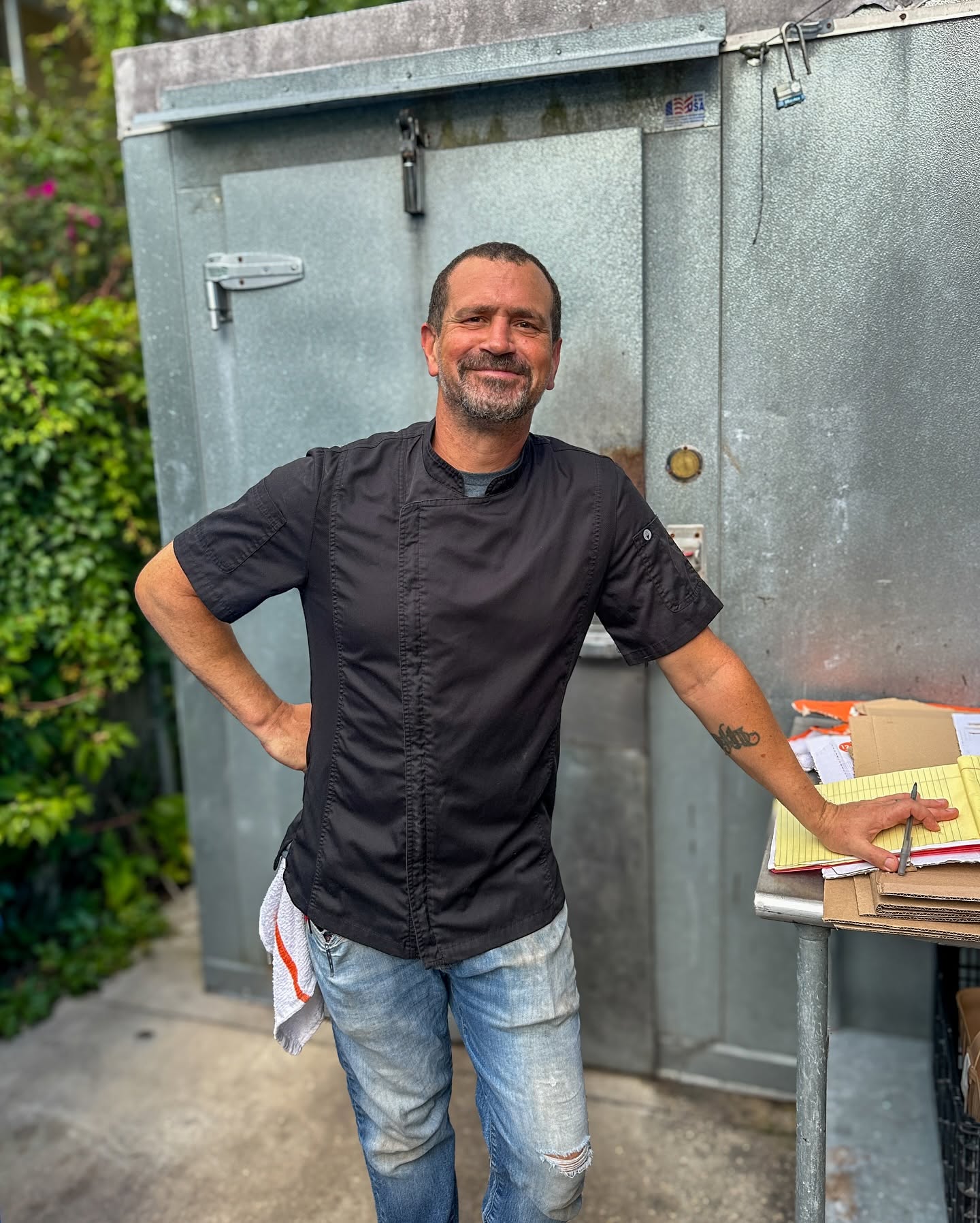 A chef in a black jacket smiling outside the kitchen door