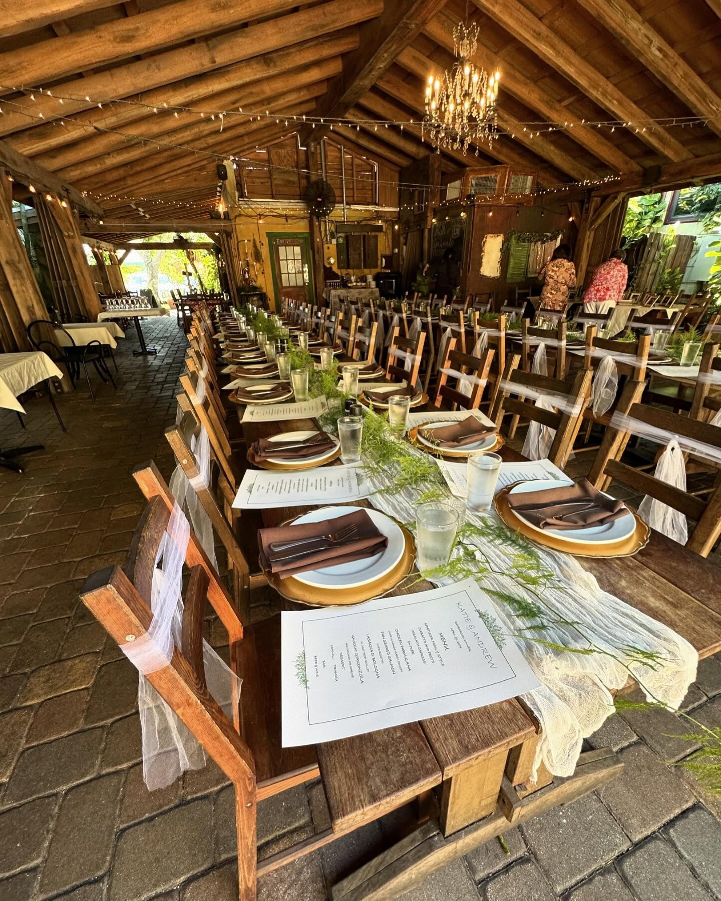 A long banquet table set with gold chargers and greenery under the timber roof of the covered patio